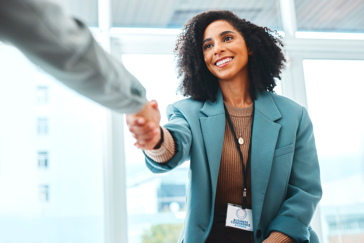 A imagem mostra uma mulher apertando a mão de outra pessoa em um amiente corporativo.